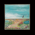 A square framed landscape painting featuring a winding dirt path through a colorful field of flowers under a teal-blue sky with a small bird in flight, displayed on a plain wall.